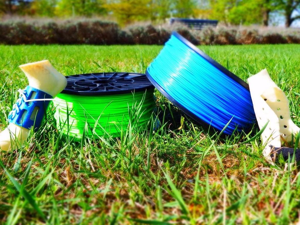 Grüne und blaue Filamentrollen neben weißen 3D-gedruckten Orthesenteilen auf einer Wiese